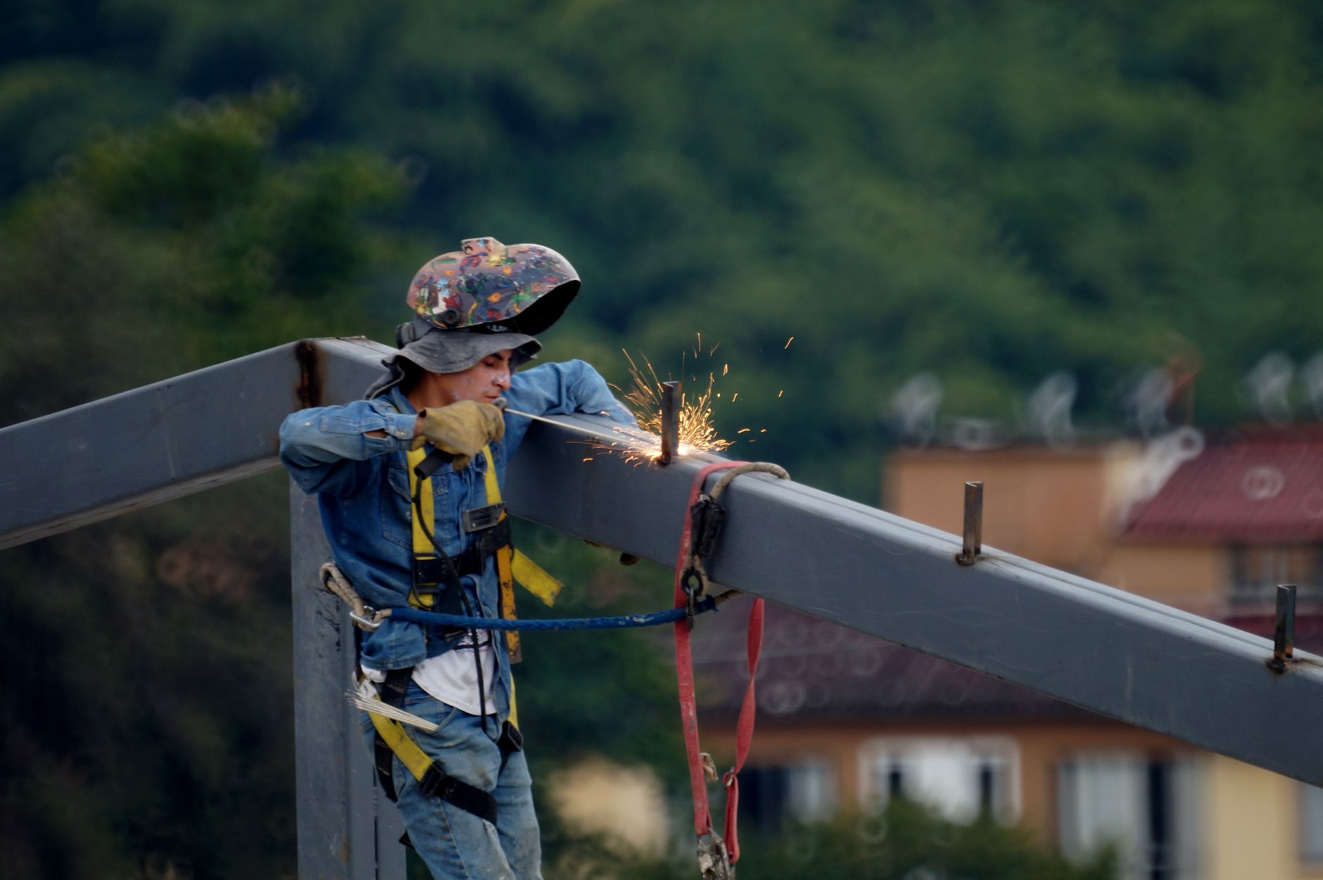 A woman in welding gear directs a torch toward a steel workpiece inside a metal fabrication shop, sparks scattering across the table.
